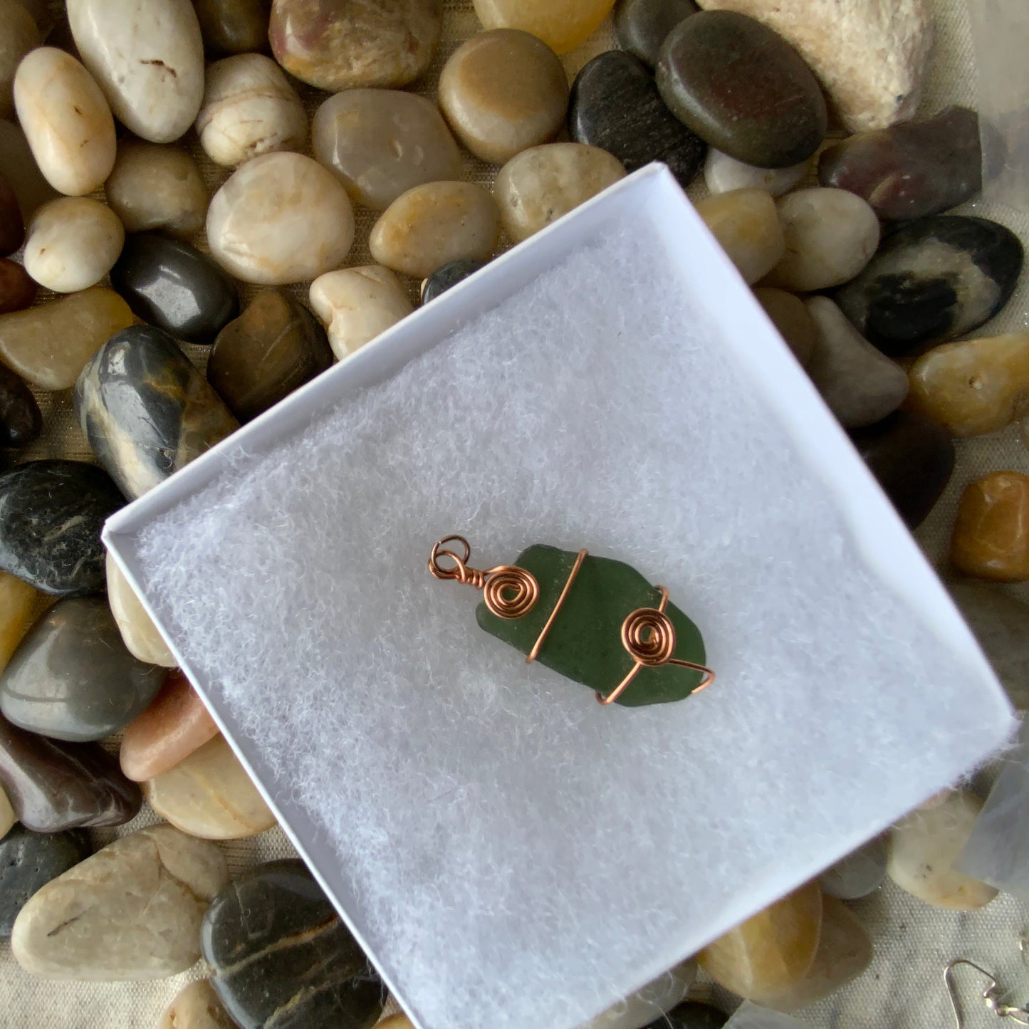 Green leaf-shaped pendant with copper wire wrapping on a white box against a background of pebbles.